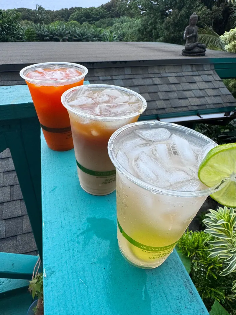Thai iced tea, Thai iced coffee and lemonade with lime on a teal rail, with Hāna rain forest behind