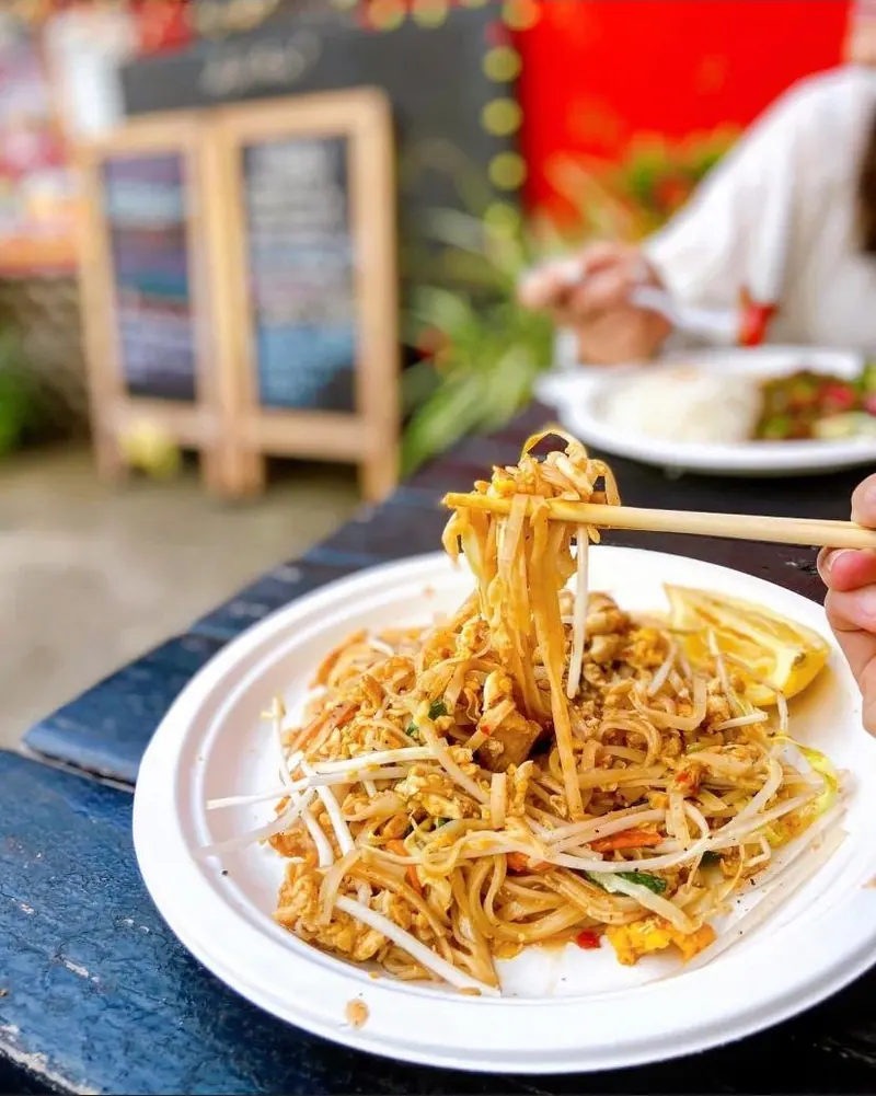 Pad thai lifted with chopsticks at a picnic table, with the chalkboard menus and the red truck behind