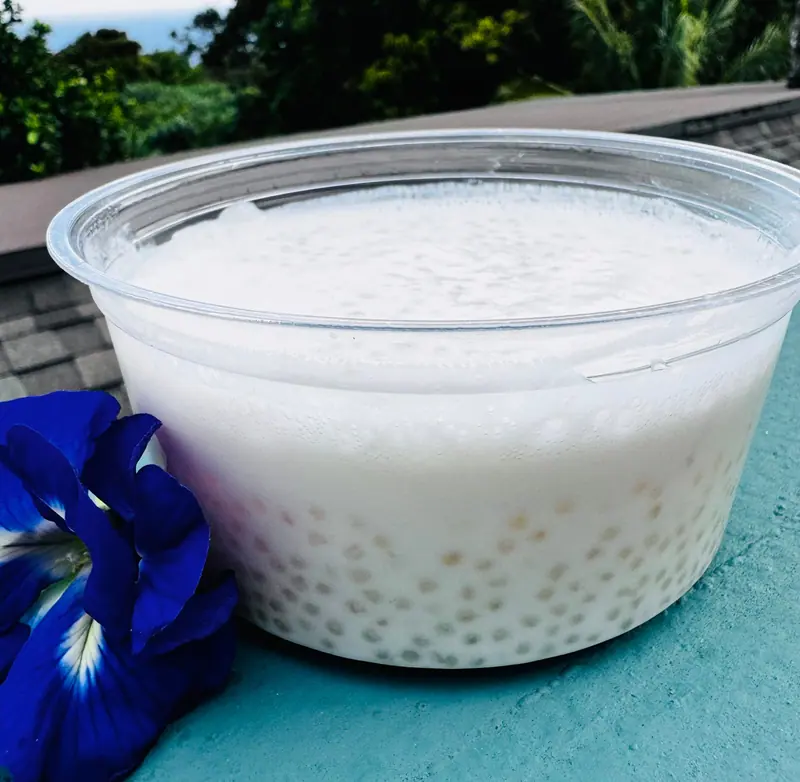 Coconut tapioca pudding in a clear cup next to a blue butterfly pea flower, with Hāna rain forest behind