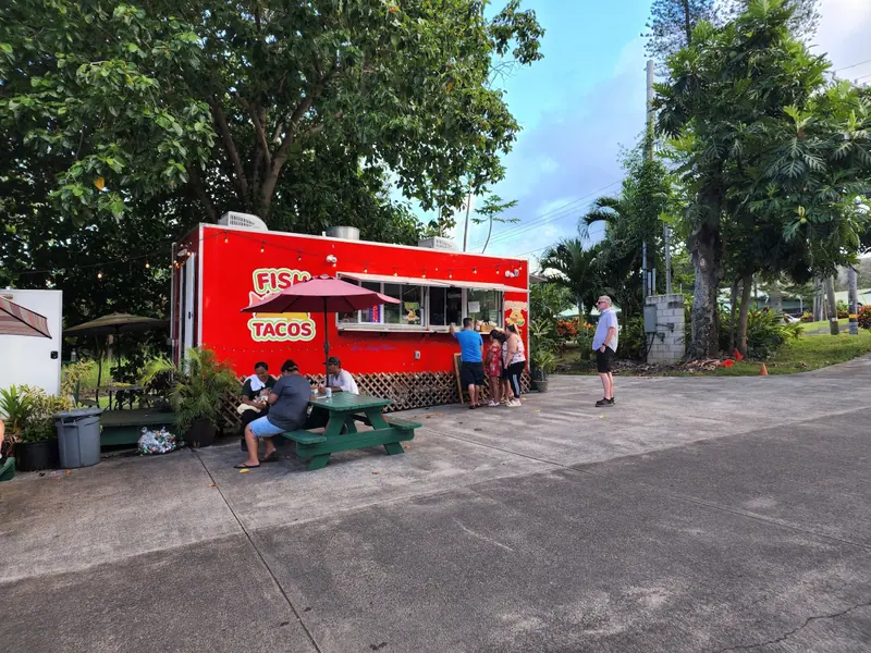 The red taco truck parked in the lot in Hāna town, with picnic tables under big shade trees